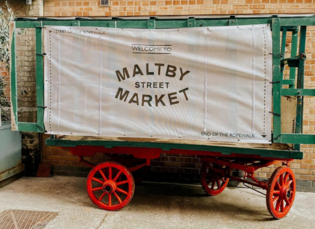 The image shows an old wooden trailer painted green with red spoke wheels. The trailer is being used to advertise Maltby Street Market as there is a white banner draped across it reading 'Welcome to Maltby Street Market'. The trailer is positioned against a yellow brick wall. 
