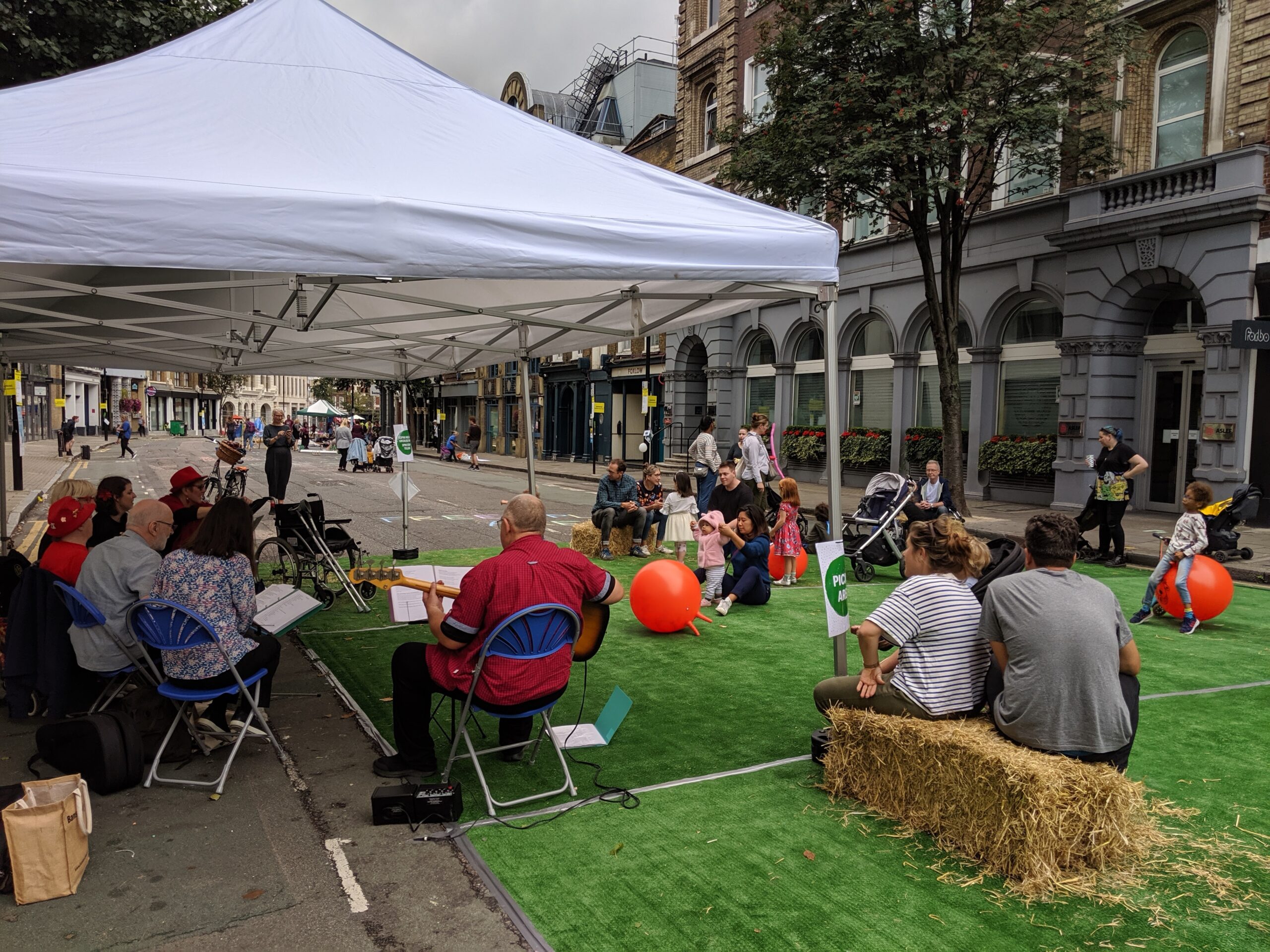 The image shows a street in North London which has been reappropriated as part of the city's 2019 car free day celebrations. In the foreground, synthetic turf covers a main road with families sat on straw bales listening as a band play. The band are sat down underneath a white gazebo. In the distance are other car free day events. 