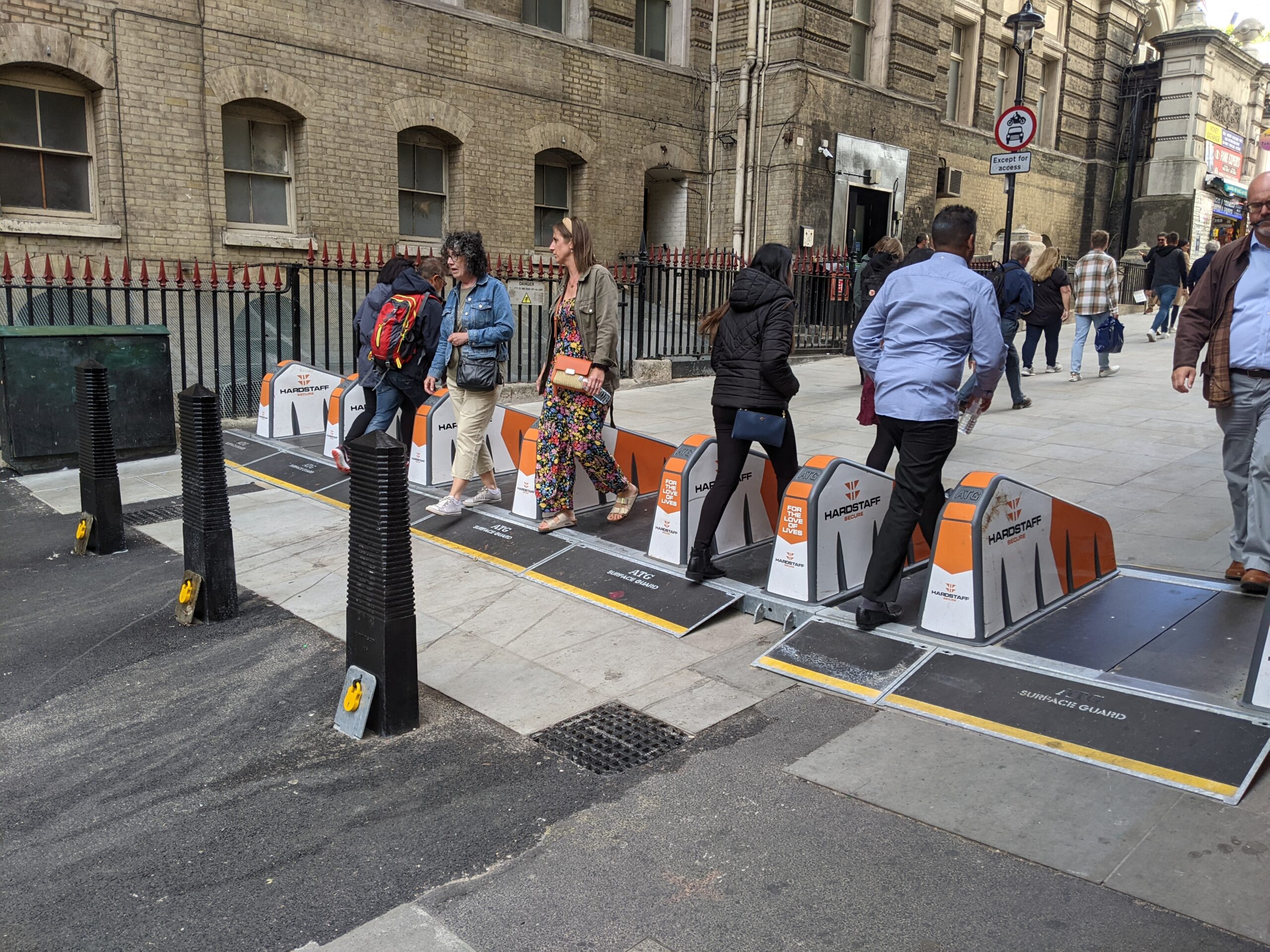 The image shows a London street that has been pedestrianised via bollards and a temporary barrier running the width of the street. People walk through the barrier, while in the background a road sign says 'except for access'. 