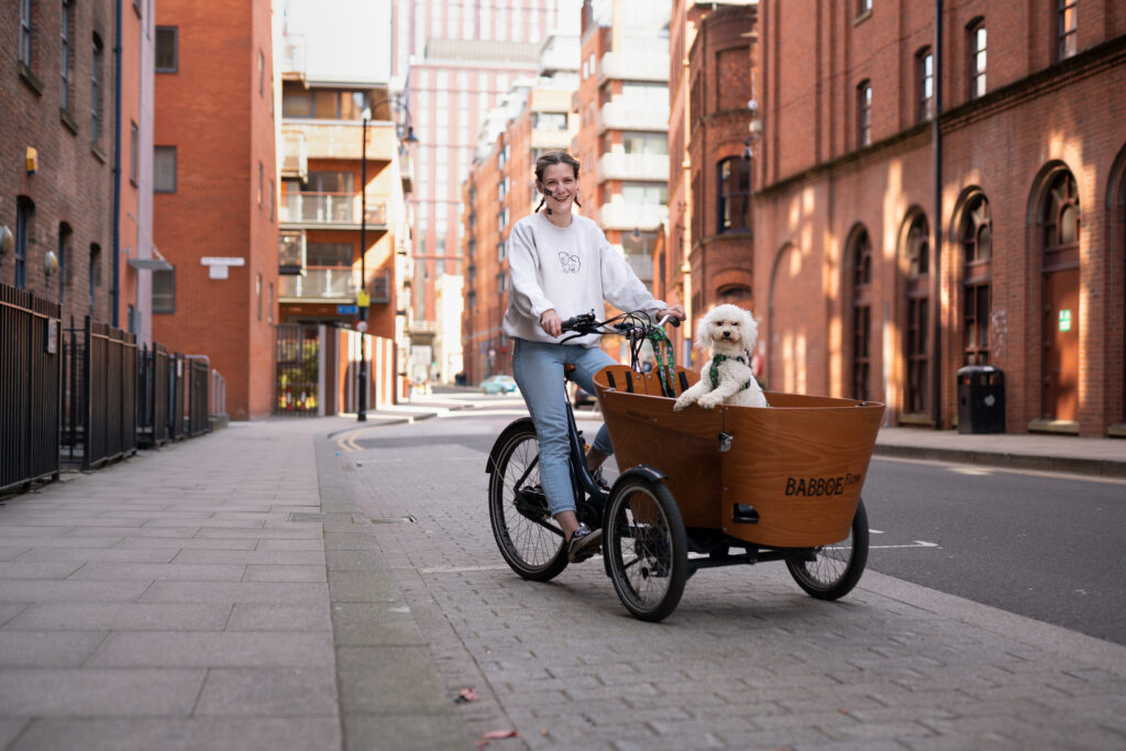 Harrie cycles her Babboe cargo trike amongst the midrise brick buildings of Ancoats, accompanied by her white Cavapoo Frida who sits in the cargo area.