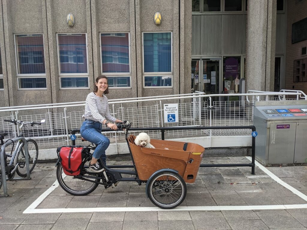 Harrie sits smiling on the seat of her Babboe cargo trike in a cycle parking space at the entrance to a Manchester University building. Her white Cavapoo Frida sits in the cargo area. The space is large and has a long black railing to attach cycles to. The railing has a sign that says "Reserved for disabled cyclists" with a standard blue wheelchair symbol beneath the text. Two blue wheelchair symbols are also present at each end of the railing.