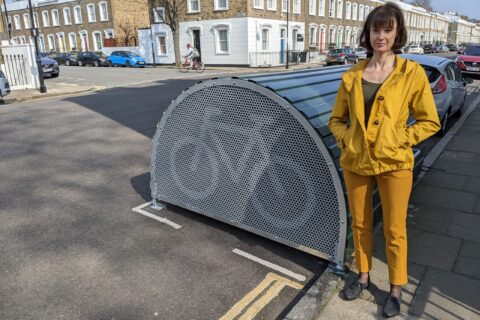 The image shows a lady standing alongside a cycle hangar on a residential street in the London borough of Islington. The lady stands in the foreground of the image and wears yellow trousers and a yellow coat. She faces the camera and has her hands in her pockets. In the background are terraced houses and parked cars. It is a sunny day and the shadow of the building to her right has been cast over the road directly behind her.