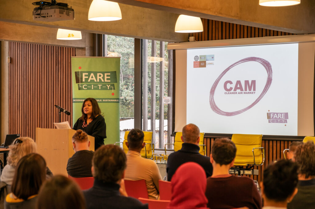 A woman stands behind a wooden lectern and is addressing an audience. She wears a black top and speaks into a microphone. In the background is a green roller banner with 'Fare City' across it, and a projector screen with the project logo and project partner logos displayed. In the foreground are the backs of audience members.