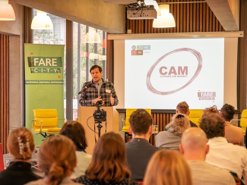 A man stands behind a wooden lectern and is addressing an audience. He wears a check shirt and speaks into a microphone. In the background is a green roller banner with 'Fare City' across it, and a projector screen with the project logo and project partner logos displayed. In the foreground are the backs of audience members.